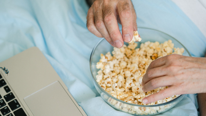 Des mains atteignent du pop-corn dans un bol en verre à côté d'un ordinateur portable, préparé pour une soirée cinéma confortable à la maison.