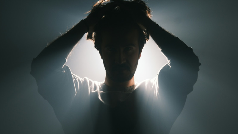 A powerful and moody portrait of a man holding his head, illuminated by an intense backlight that creates dramatic rays of light. The contrast between darkness and illumination enhances the emotions of stress, contemplation, or inner conflict. The cinemat