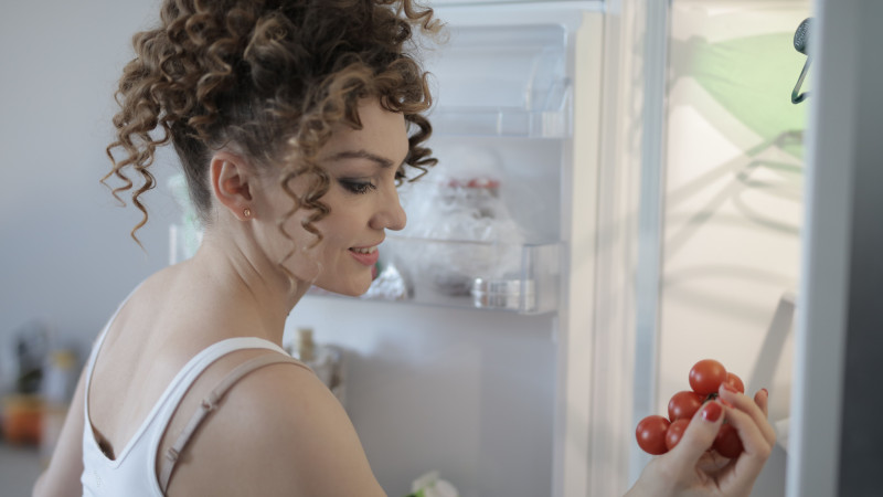 Une femme adulte joyeuse choisissant des tomates fraîches dans le réfrigérateur à la maison.