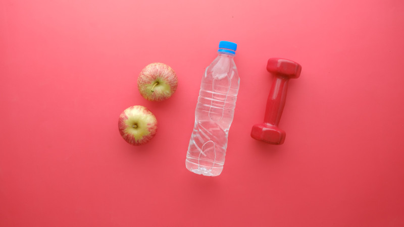 Vue de dessus d'une bouteille d'eau, de pommes et d'un haltère sur un fond rouge vif. Parfait pour des thèmes de fitness et de bien-être.