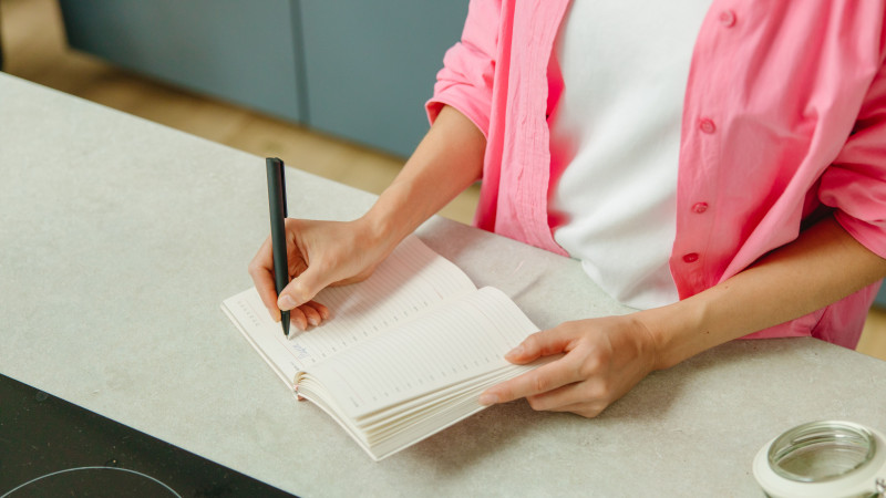 Une femme en chemise rose écrivant dans un carnet sur un plan de travail de cuisine avec un stylo.