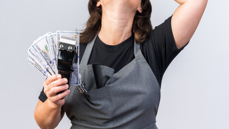Coiffeuse caucasienne adulte mécontente en uniforme mettant la main sur le front et tenant de l'argent avec une tondeuse à cheveux isolée sur fond blanc avec espace de copie