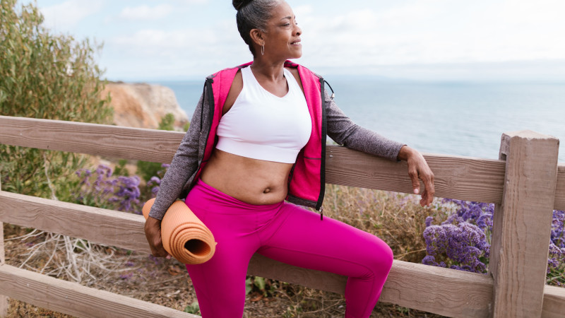 Femme senior confiante en tenue de sport tenant un tapis de yoga, profitant d'une vue pittoresque en plein air sur l'océan.