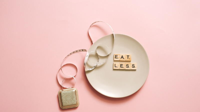 Une image motivante présentant une assiette avec des tuiles de scrabble épelant 'Mange Moins' sur un fond rose.